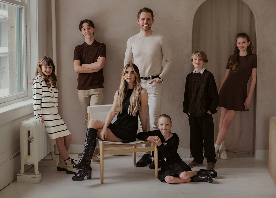 A family of seven standing and sitting in a modern, neutral-toned room, dressed in coordinated outfits, smiling and looking at the camera.
