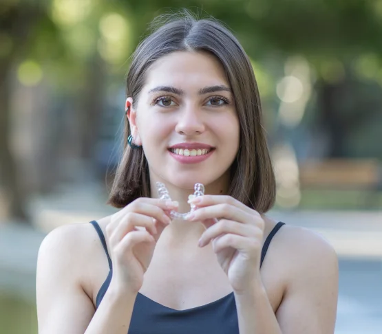 Smiling woman holding a clear aligner tray in front of her, standing outdoors with a blurred park in the background